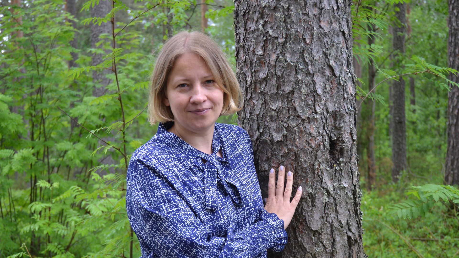 Lähikuvassa siniseen paitaa pukeutunut henkilö nojaa mäntyyn ja toinen käsi koskettaa puuta. 